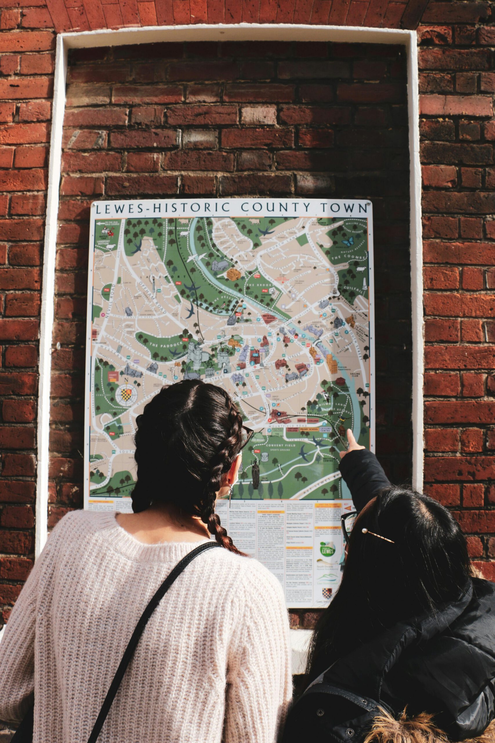 Two women study the Lewes historical town map mounted on a brick wall, pointing and discussing routes.